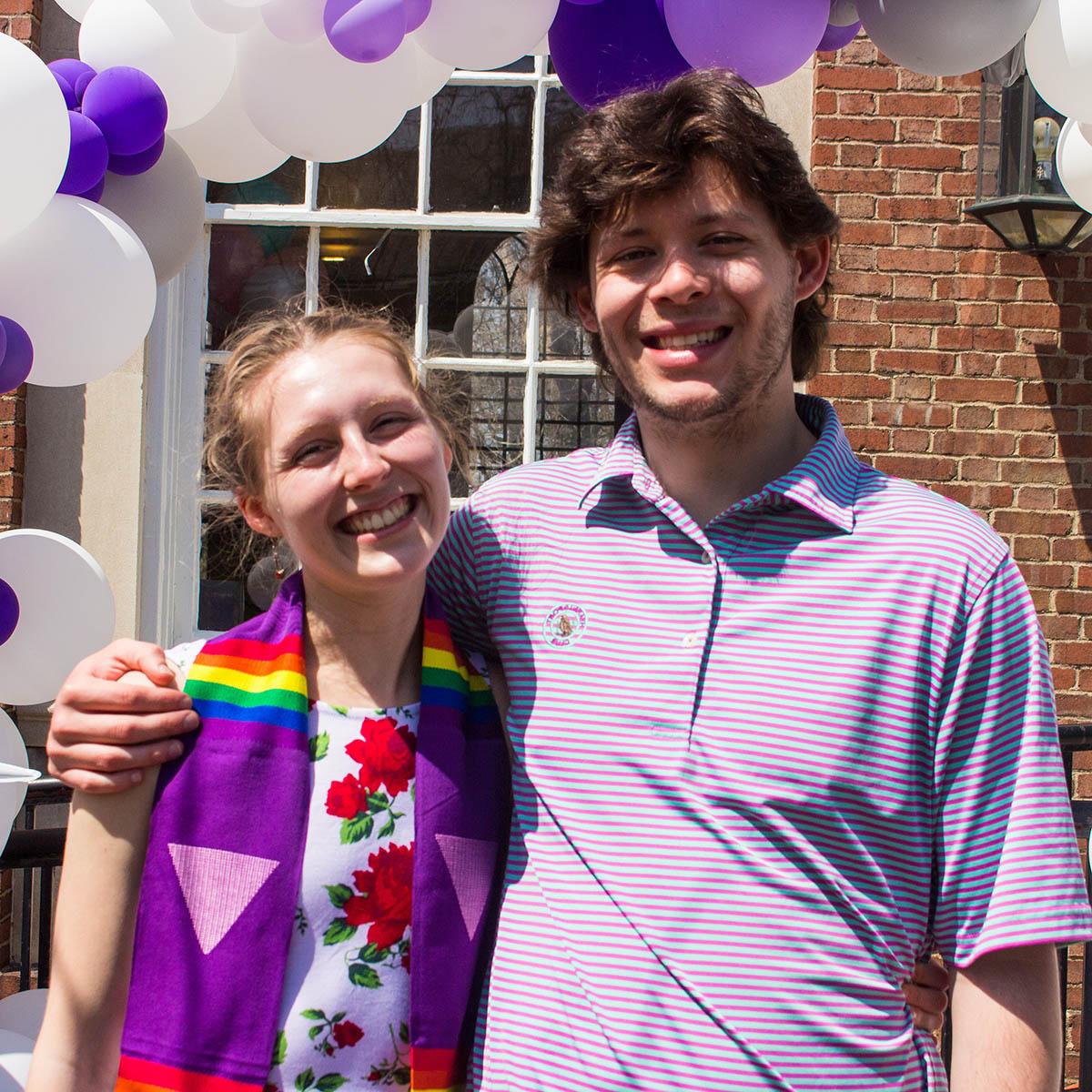 Photo of a young woman wearing a rainbow graduation stole, posing with a young man and smiling with purple and white balloons in the background