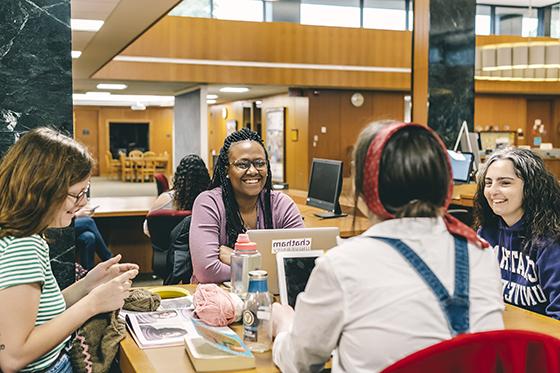 Photo of a group of Chatham University students sitting around a study table at Jennie King Mellon Library smiling and studying together. 